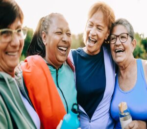 Four senior women laughing and embracing each other outdoors.