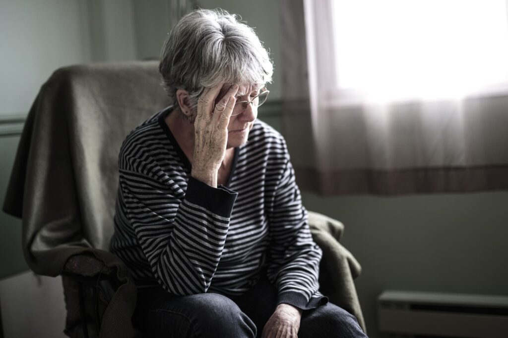 Elderly woman sitting in a chair with her hand on her forehead, looking distressed.