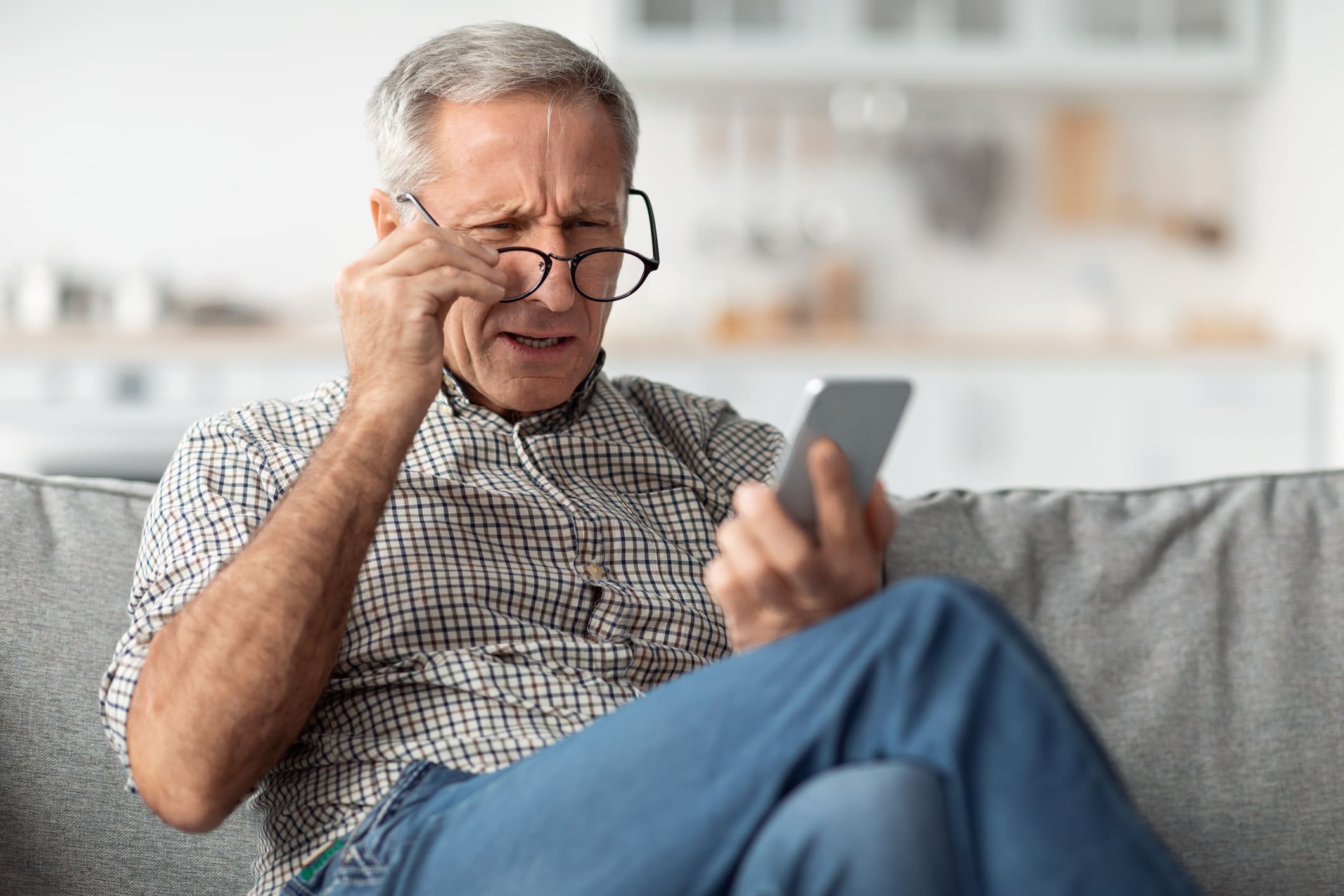 Elderly man sitting on a sofa, squinting and adjusting his glasses while looking at a smartphone.