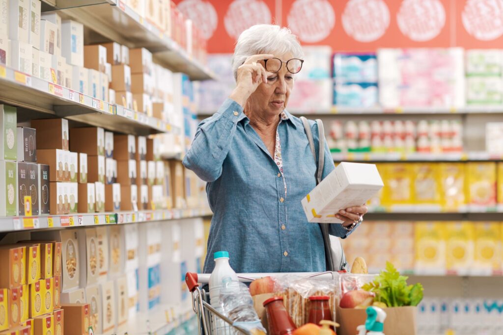 Senior woman with glasses carefully reading a product label while shopping for groceries, highlighting the importance of vision health in daily tasks.