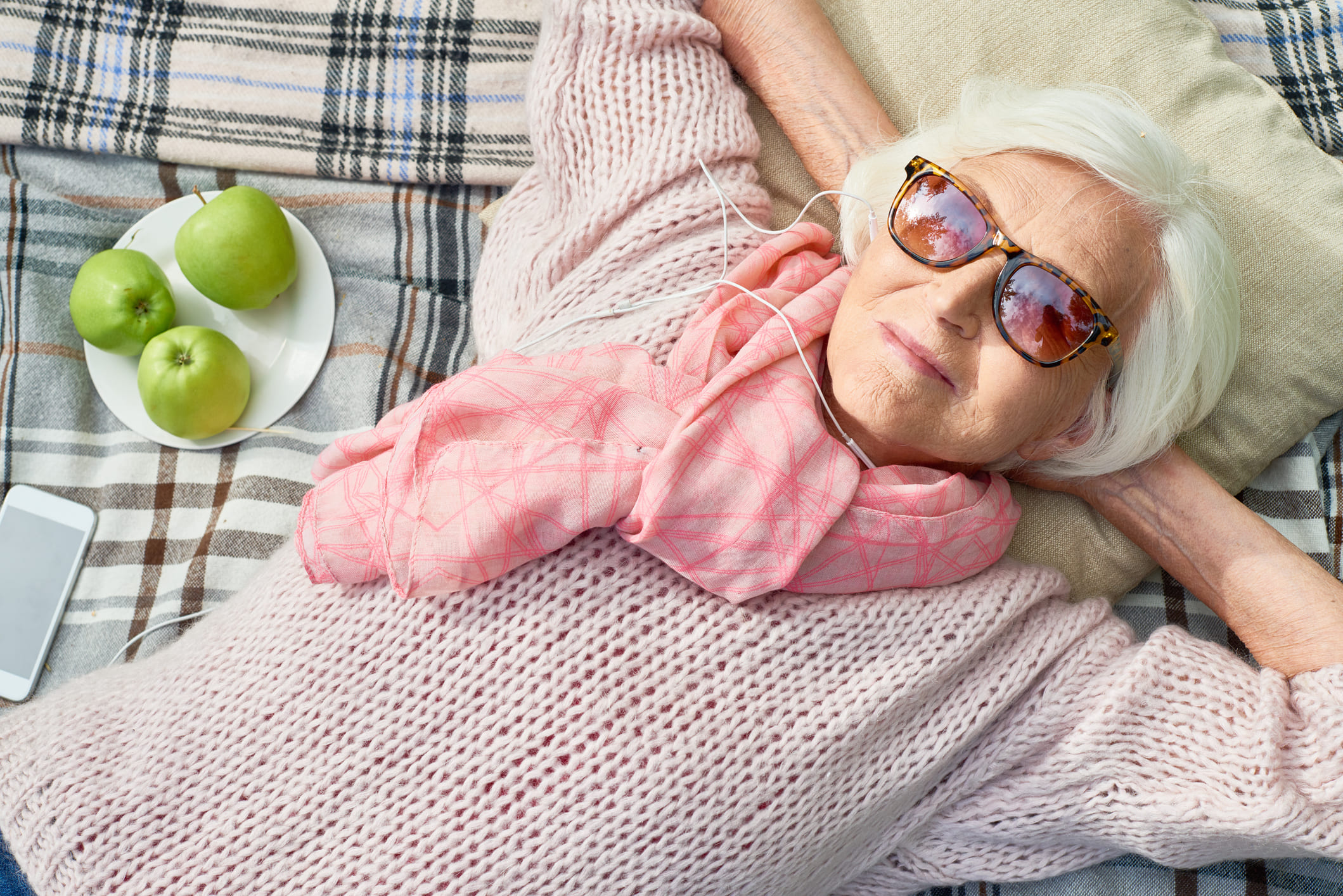 Senior woman relaxing outdoors with sunglasses on, lying on a blanket next to a plate of green apples.