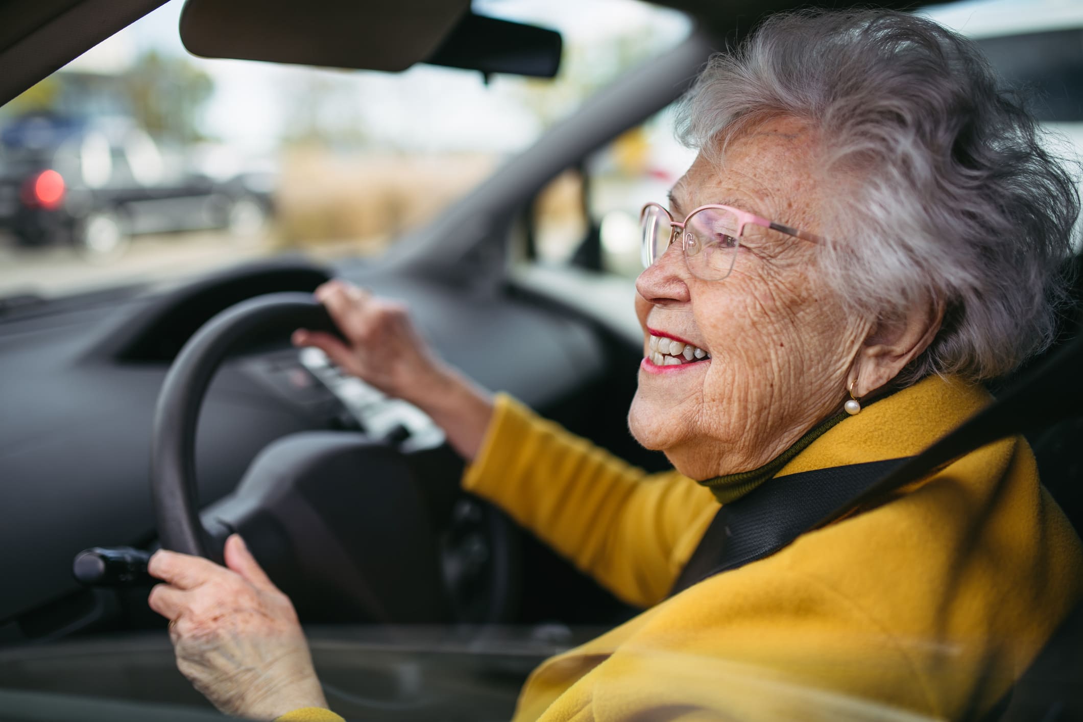 Smiling elderly woman with glasses confidently driving a car, showcasing independence and the ability to drive safely despite living with macular degeneration.