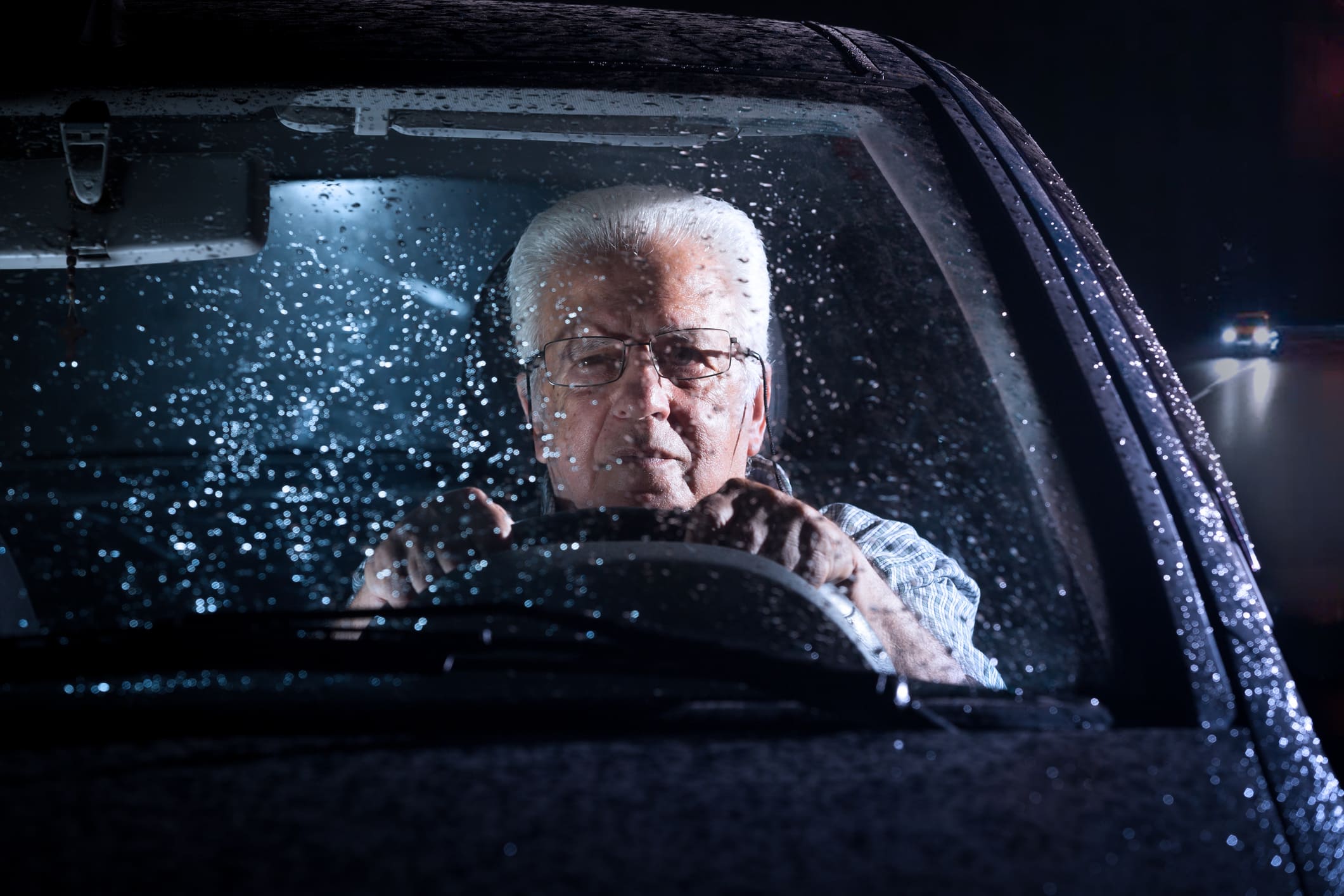 Older man driving a car at night in the rain, struggling to see clearly through the windshield, representing the challenges of night blindness while driving.