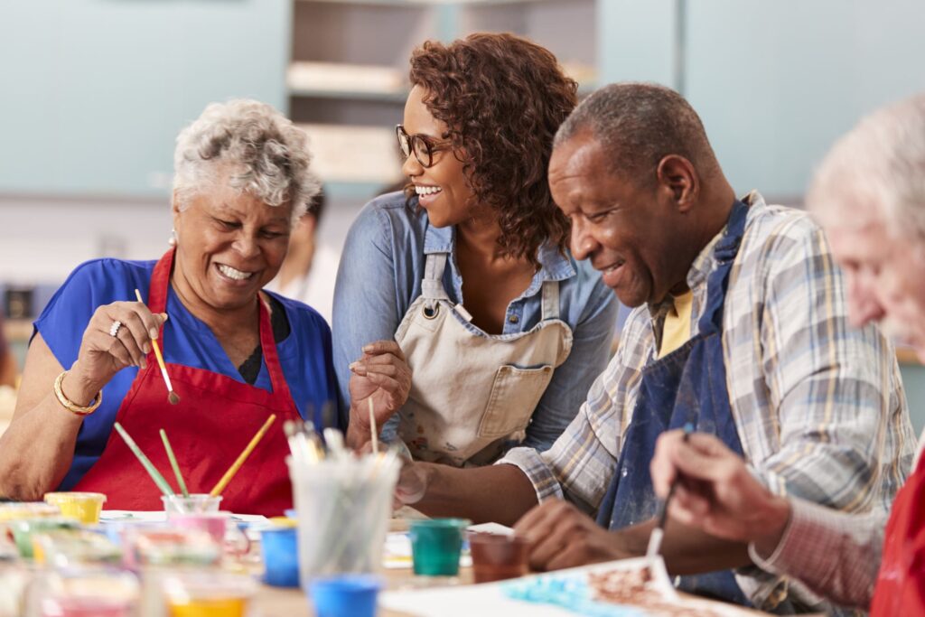 Group of older adults joyfully painting together, assisted by a younger caregiver in an art class setting.