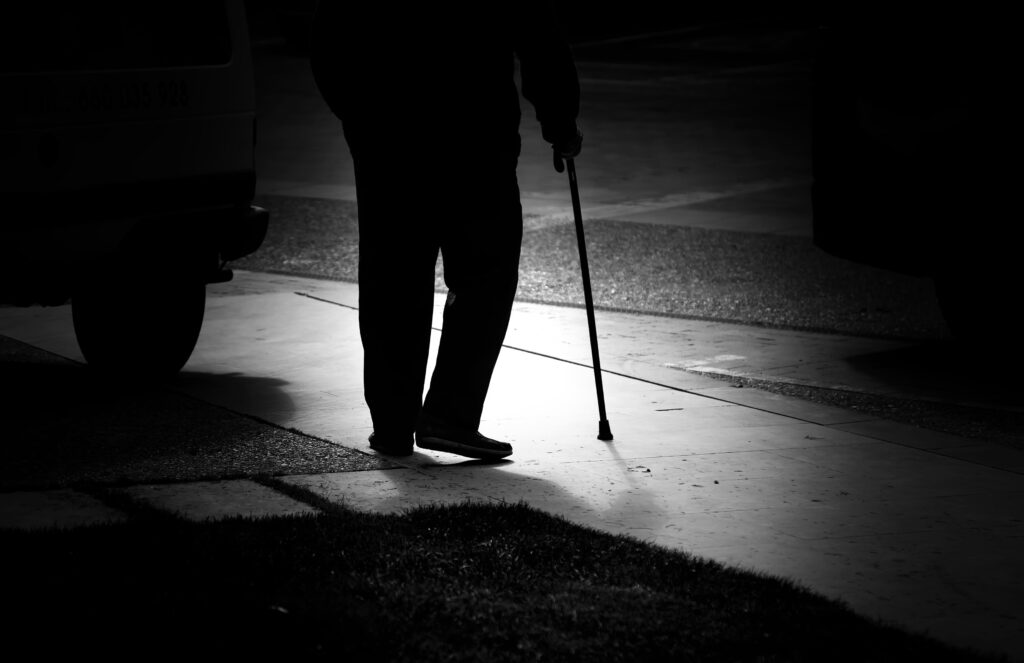 Silhouette of an elderly person walking with a cane at night on a dimly lit street, representing challenges faced due to night blindness.