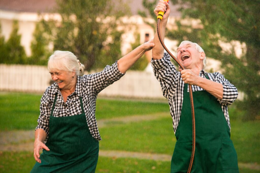 An older couple wearing matching checkered shirts and green aprons laughing and playfully spraying water with a garden hose on a sunny day.