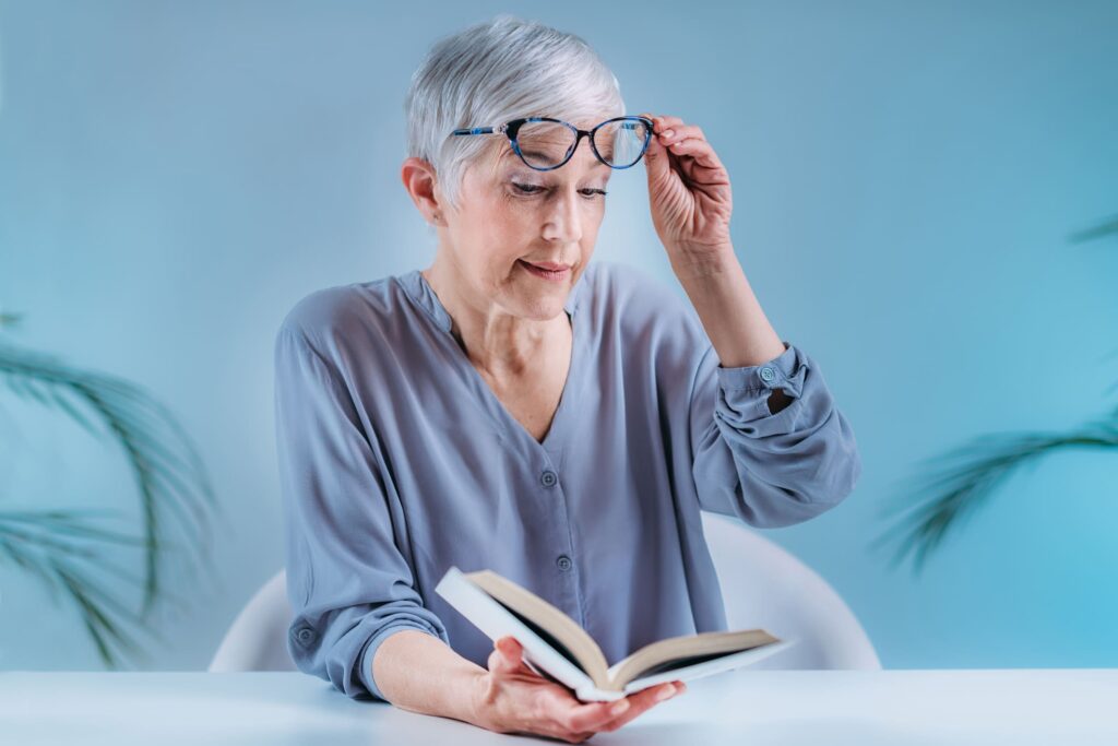 An older woman with short gray hair adjusts her glasses while reading a book, reflecting the impact of age-related macular degeneration (AMD) on daily activities.