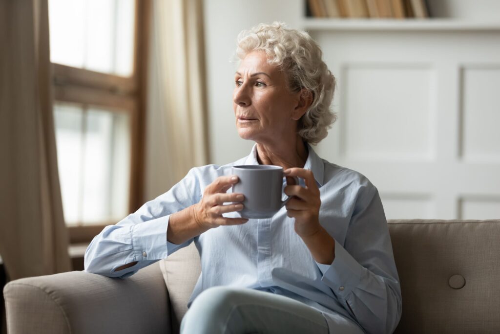 A senior woman with gray hair sitting on a couch, holding a cup, reflecting on the emotional impacts of AMD disease progression.