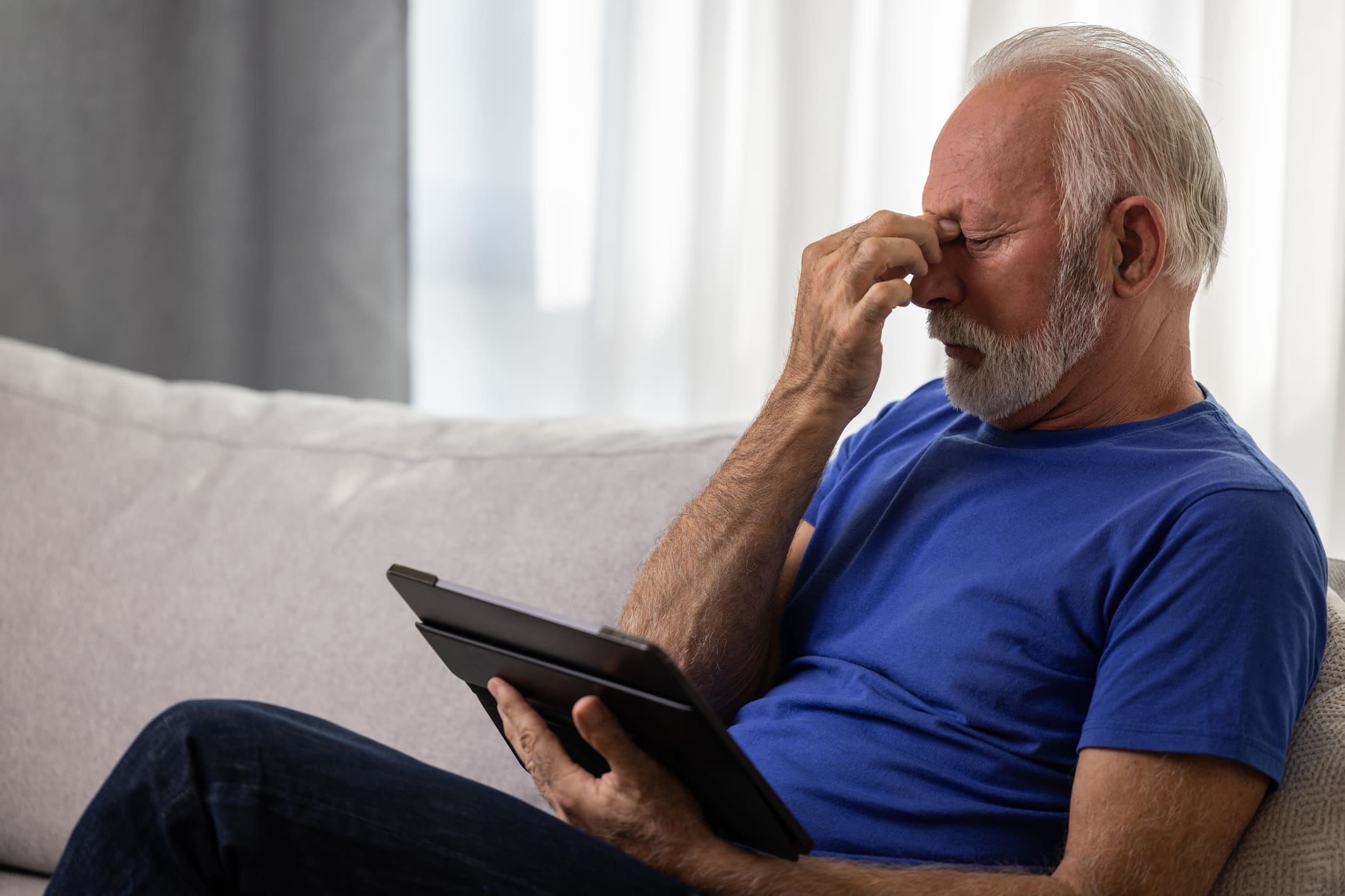 A senior man with gray hair and a beard, sitting on a couch, rubbing his eyes in frustration while holding a tablet, reflecting on the challenges of AMD disease progression.