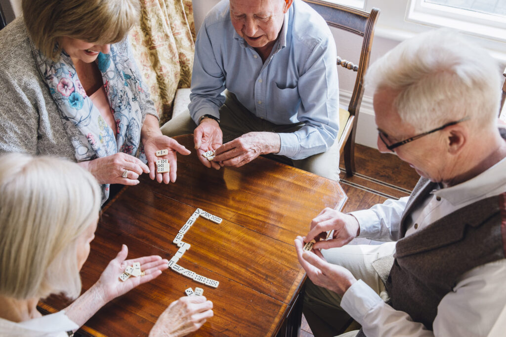 Four older adults playing a game of dominoes around a wooden table. They are focused on their tiles, with some of the pieces placed on the table forming a line.