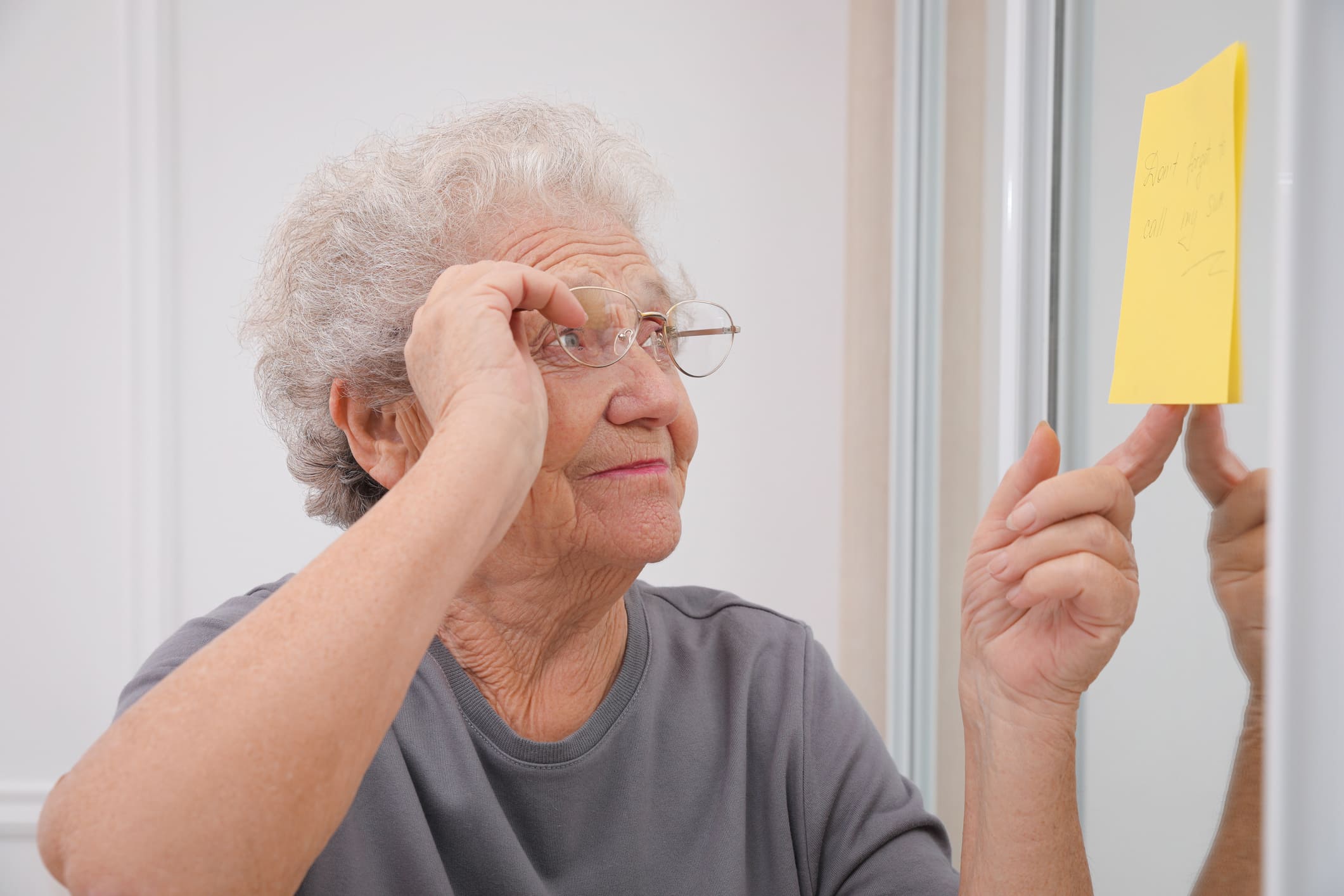 An elderly woman with short gray hair adjusts her glasses while looking at a yellow sticky note on a mirror that reads "Short term memory, call my son."